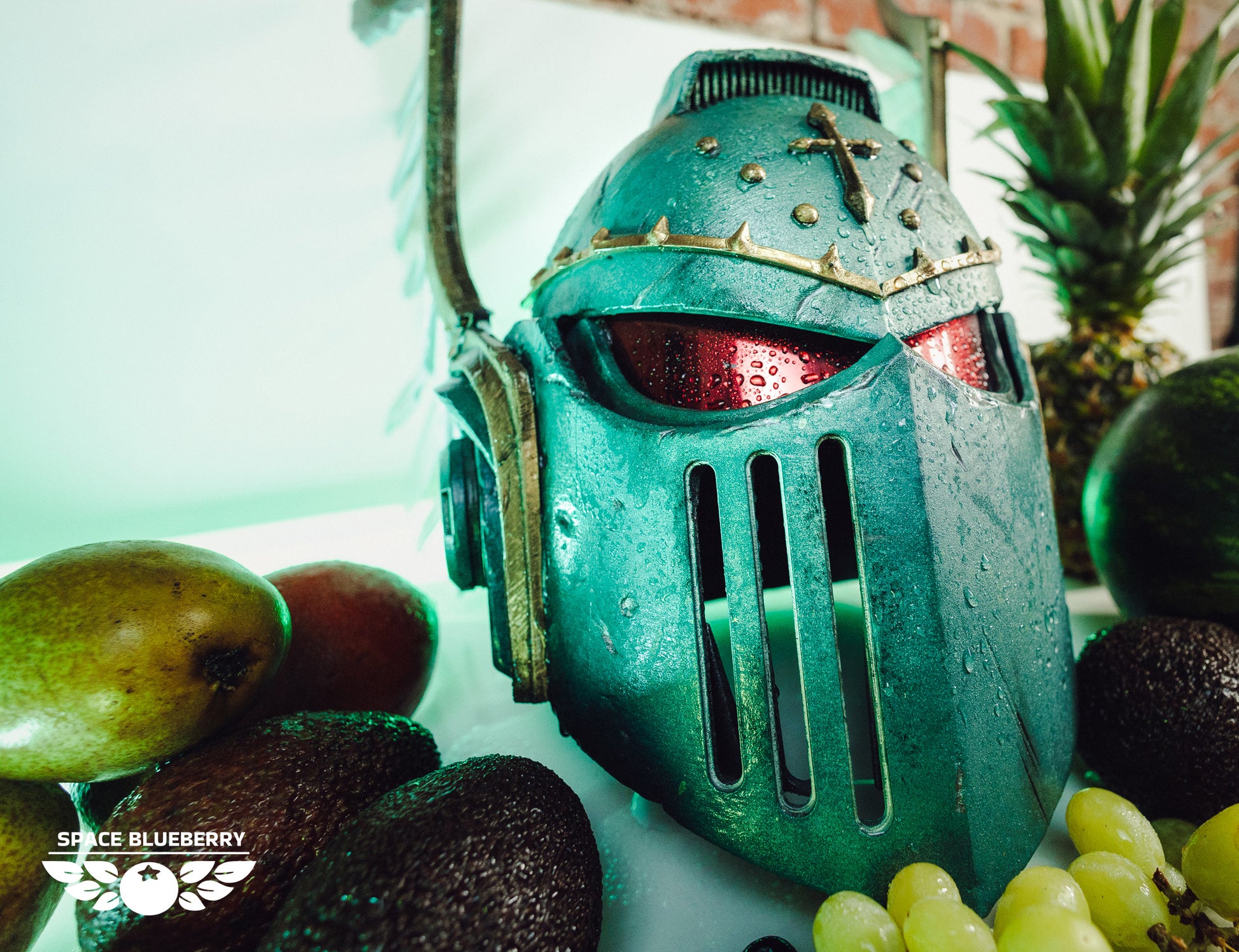 Green angels helmet with red eyes vizor surrounded by fruits on a white surface with gold and white wings with feathers.